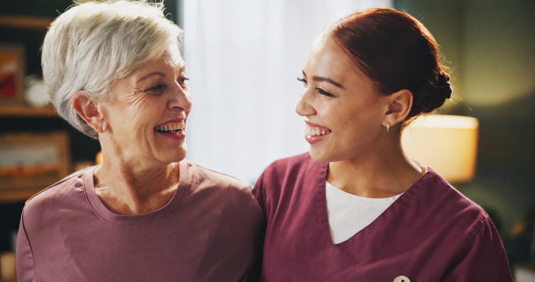 friendly caregiver and smiling elderly woman shoulder to shoulder share a laugh